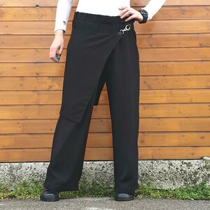 May include: Black wrap-style pants with a wide leg design. The pants feature a unique wrap detail secured with a silver-tone metal ring. The model is wearing a white long-sleeve shirt and black shoes. The background is a wooden wall.