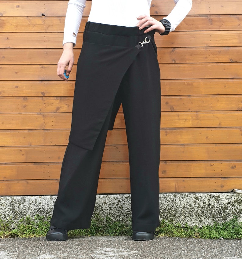 May include: Black wrap-style pants with a wide leg design. The pants feature a unique wrap detail secured with a silver-tone metal ring. The model is wearing a white long-sleeve shirt and black shoes. The background is a wooden wall.