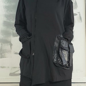 Op de afbeelding: Zwarte hoodie met een lange rits en een groot zwart nylon zakje aan de zijkant.