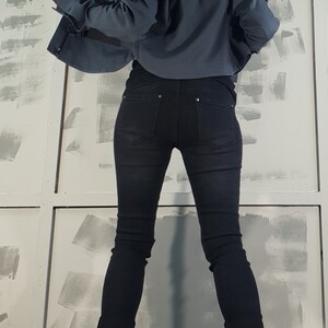 May include: A woman wearing a dark blue cropped jacket with black jeans and black ankle boots. The jacket has a zipper and a buckle detail. The jeans are skinny and the boots have laces.