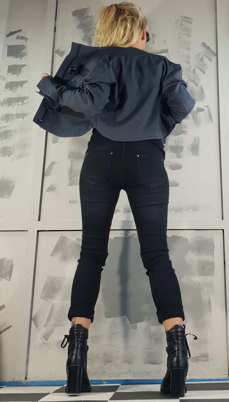 May include: A woman wearing a dark blue cropped jacket with black jeans and black ankle boots. The jacket has a zipper and a buckle detail. The jeans are skinny and the boots have laces.
