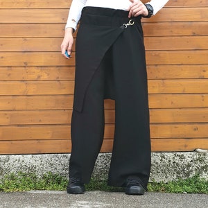 May include: Black wide-leg pants with a wrap-style front and a decorative gold-tone clasp. The pants have a high waist and a relaxed fit. The model is wearing black shoes and a white long-sleeve shirt.