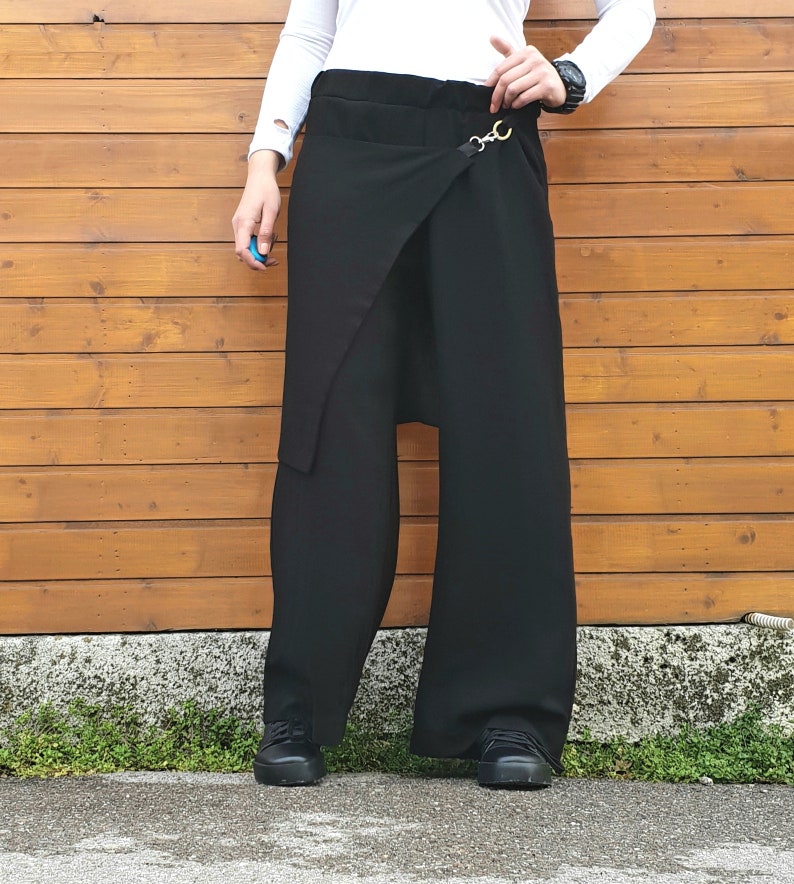 May include: Black wide-leg pants with a wrap-style front and a decorative gold-tone clasp. The pants have a high waist and a relaxed fit. The model is wearing black shoes and a white long-sleeve shirt.
