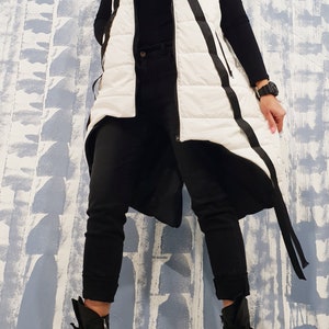 May include: A white puffer vest with black trim and a long, asymmetrical hemline. The vest has a zipper closure and a hood. The model is wearing black pants and black boots.