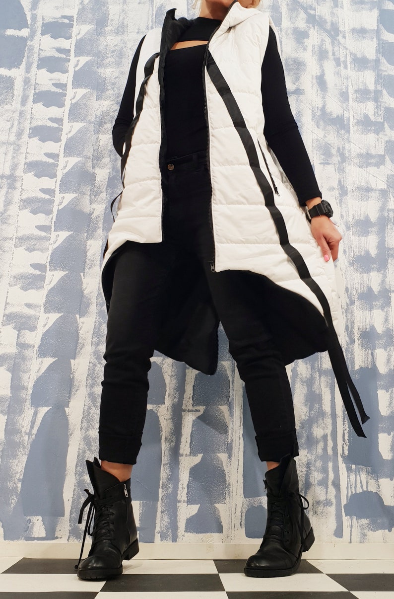 May include: A white puffer vest with black trim and a long, asymmetrical hemline. The vest has a zipper closure and a hood. The model is wearing black pants and black boots.