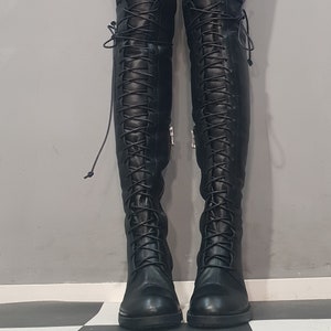 May include: A pair of black, over-the-knee boots with a lace-up design. The boots have a round toe and a small heel. The laces run up the front of the boots. The boots are made of a smooth material and have a zipper on the side.