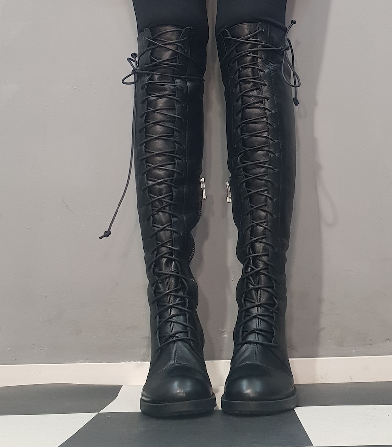 May include: A pair of black, over-the-knee boots with a lace-up design. The boots have a round toe and a small heel. The laces run up the front of the boots. The boots are made of a smooth material and have a zipper on the side.