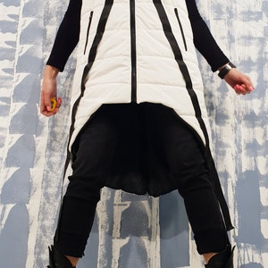 May include: A white puffer vest with black straps and a zipper closure. The vest is long and has a high neckline. The model is wearing black pants and black boots.