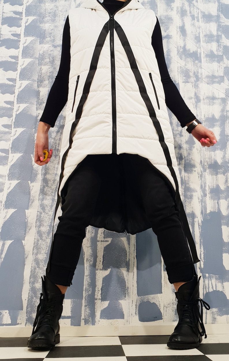 May include: A white puffer vest with black straps and a zipper closure. The vest is long and has a high neckline. The model is wearing black pants and black boots.