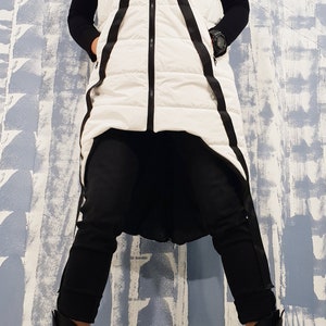 May include: A white puffer vest with black straps and a zipper closure. The vest is long and has a high collar. The person is wearing black pants and black boots.