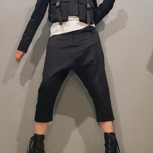May include: A black cropped jacket with a zipper closure and multiple straps and buckles. The jacket is paired with black pants and black ankle boots.
