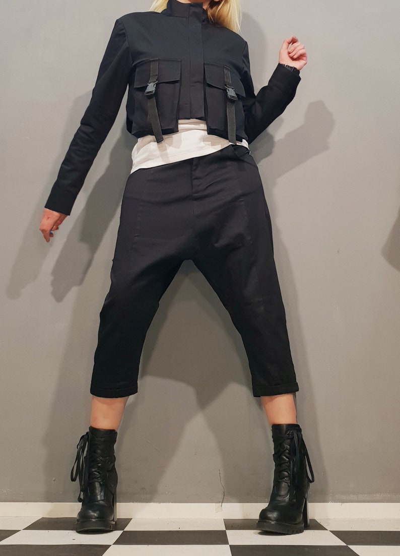 May include: A black cropped jacket with a zipper closure and multiple straps and buckles. The jacket is paired with black pants and black ankle boots.