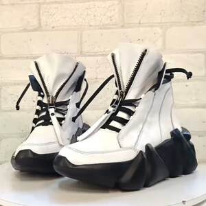 May include: A pair of white high-top sneakers with black accents. The sneakers feature a zipper closure, black laces, and a unique black, geometric-shaped sole. The shoes are displayed on a white surface against a white brick wall.