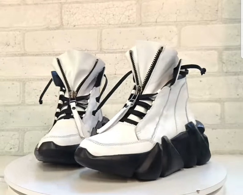 May include: A pair of white high-top sneakers with black accents. The sneakers feature a zipper closure, black laces, and a unique black, geometric-shaped sole. The shoes are displayed on a white surface against a white brick wall.