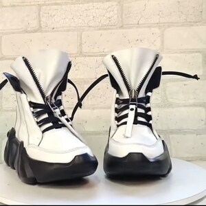 May include: A pair of white high-top boots with black accents. The boots feature a zipper closure, black laces, and a thick, black sole. The boots are set against a white brick wall.