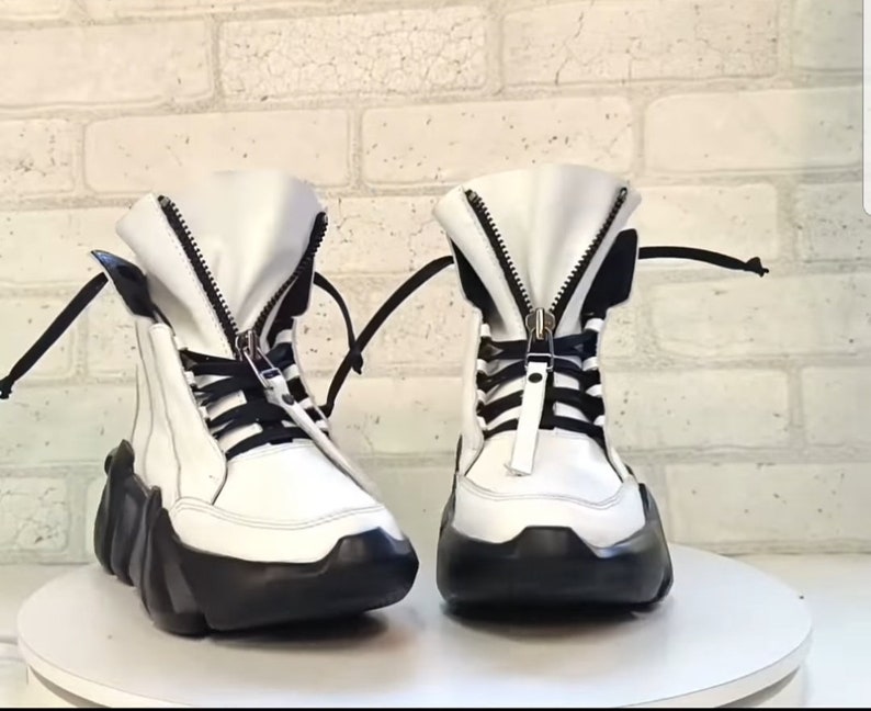 May include: A pair of white high-top boots with black accents. The boots feature a zipper closure, black laces, and a thick, black sole. The boots are set against a white brick wall.