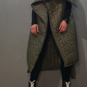 May include: A long, green quilted vest with a large, folded collar. The vest is sleeveless and has a relaxed fit. The model is wearing black trousers and black boots with white laces.