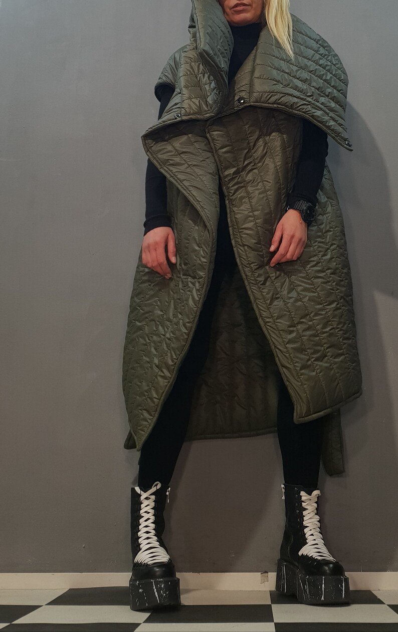 May include: A long, green quilted vest with a large, folded collar. The vest is sleeveless and has a relaxed fit. The model is wearing black trousers and black boots with white laces.