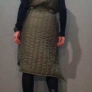 May include: A long, quilted vest with a high collar and a belt. The vest is olive green and has a slightly asymmetrical hemline.