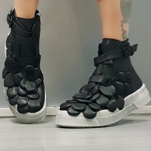May include: Black high-top sneakers with a unique design featuring 3D flower-shaped embellishments. The shoes have a white sole and a black leather upper.