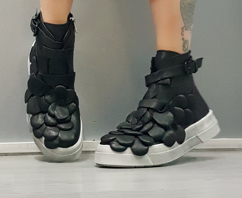 May include: Black high-top sneakers with a unique design featuring 3D flower-shaped embellishments. The shoes have a white sole and a black leather upper.