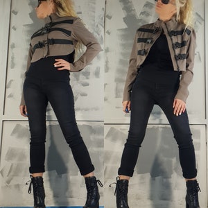 May include: A brown cropped jacket with black straps and buckles. The jacket has a high collar and long sleeves. The model is wearing black pants and black boots.