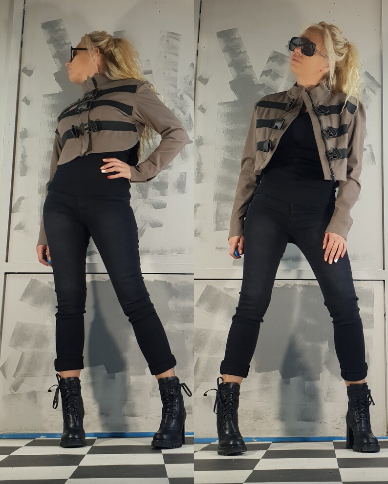 May include: A brown cropped jacket with black straps and buckles. The jacket has a high collar and long sleeves. The model is wearing black pants and black boots.