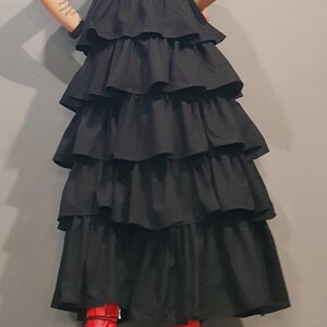 May include: A black sleeveless dress with multiple layers of ruffles. The dress is long and falls to the ankles. The model is wearing red platform boots.