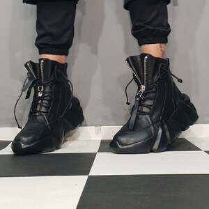 May include: Black leather ankle boots with a unique, geometric design. The boots feature a front zipper, lace-up detail, and a thick, sculpted sole. The boots are worn with black pants.
