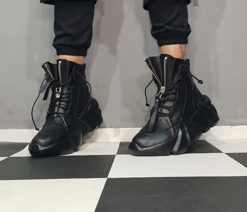 May include: Black leather ankle boots with a unique, geometric design. The boots feature a front zipper, lace-up detail, and a thick, sculpted sole. The boots are worn with black pants.