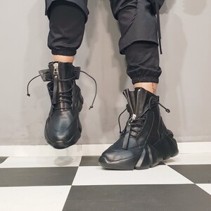 May include: Black leather high-top boots with a zipper closure and lace-up detail. The boots have a unique, chunky sole and are set against a black and white checkered floor. The boots are part of a fashion-forward footwear collection.