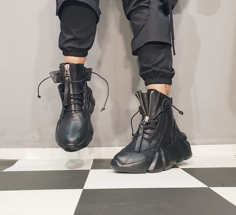May include: Black leather high-top boots with a zipper closure and lace-up detail. The boots have a unique, chunky sole and are set against a black and white checkered floor. The boots are part of a fashion-forward footwear collection.