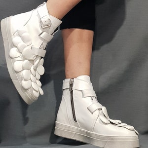 May include: White leather high-top sneakers with a unique flower-shaped design on the side. The sneakers have a zipper on the side and a buckle closure.