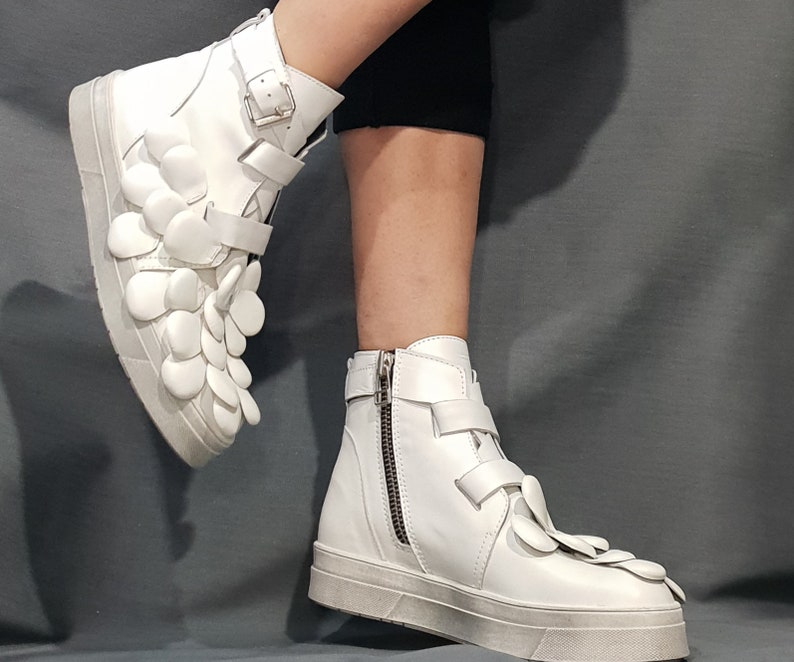May include: White leather high-top sneakers with a unique flower-shaped design on the side. The sneakers have a zipper on the side and a buckle closure.