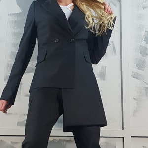 May include: A black blazer with a double-breasted closure and a long, asymmetrical hemline. The blazer is worn over a white top and black pants.