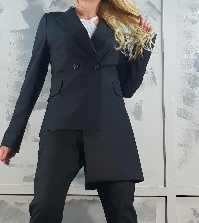 May include: A black blazer with a double-breasted closure and a long, asymmetrical hemline. The blazer is worn over a white top and black pants.