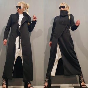 May include: A long black coat with a unique asymmetrical design, featuring a high collar and a split front that reveals white pants. The outfit is completed with black heeled sandals and sunglasses, creating a modern, edgy look.