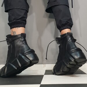 May include: Black leather ankle boots with a unique, geometric platform sole. The boots have a side zipper and a lace-up detail at the top. The boots are worn with black pants.