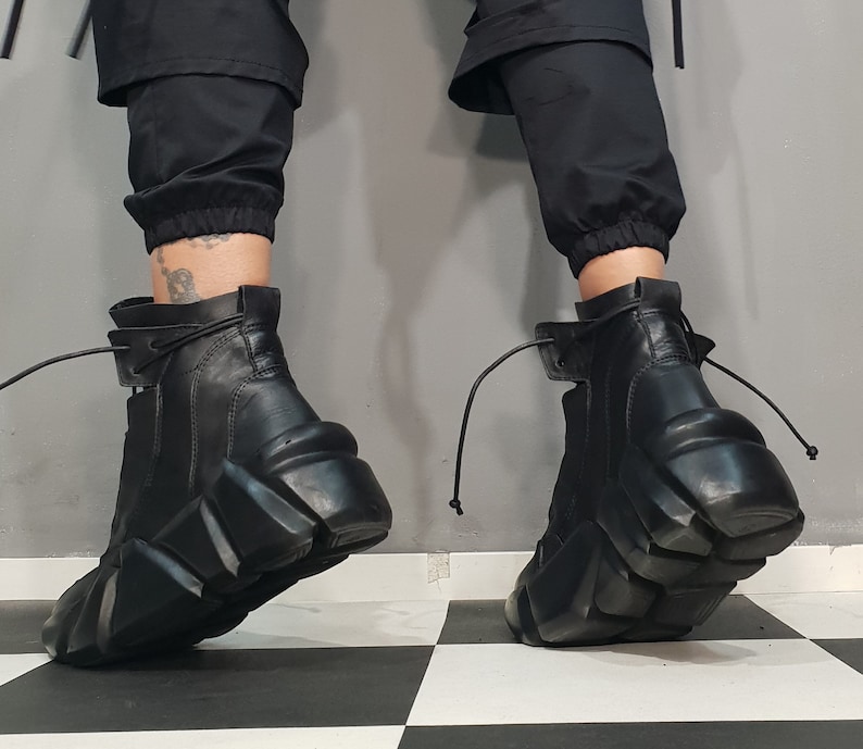 May include: Black leather ankle boots with a unique, geometric platform sole. The boots have a side zipper and a lace-up detail at the top. The boots are worn with black pants.