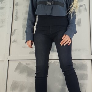 May include: A woman wearing a blue cropped jacket with black straps and buckles, black jeans, and black lace-up boots. She is wearing sunglasses and has blonde hair.