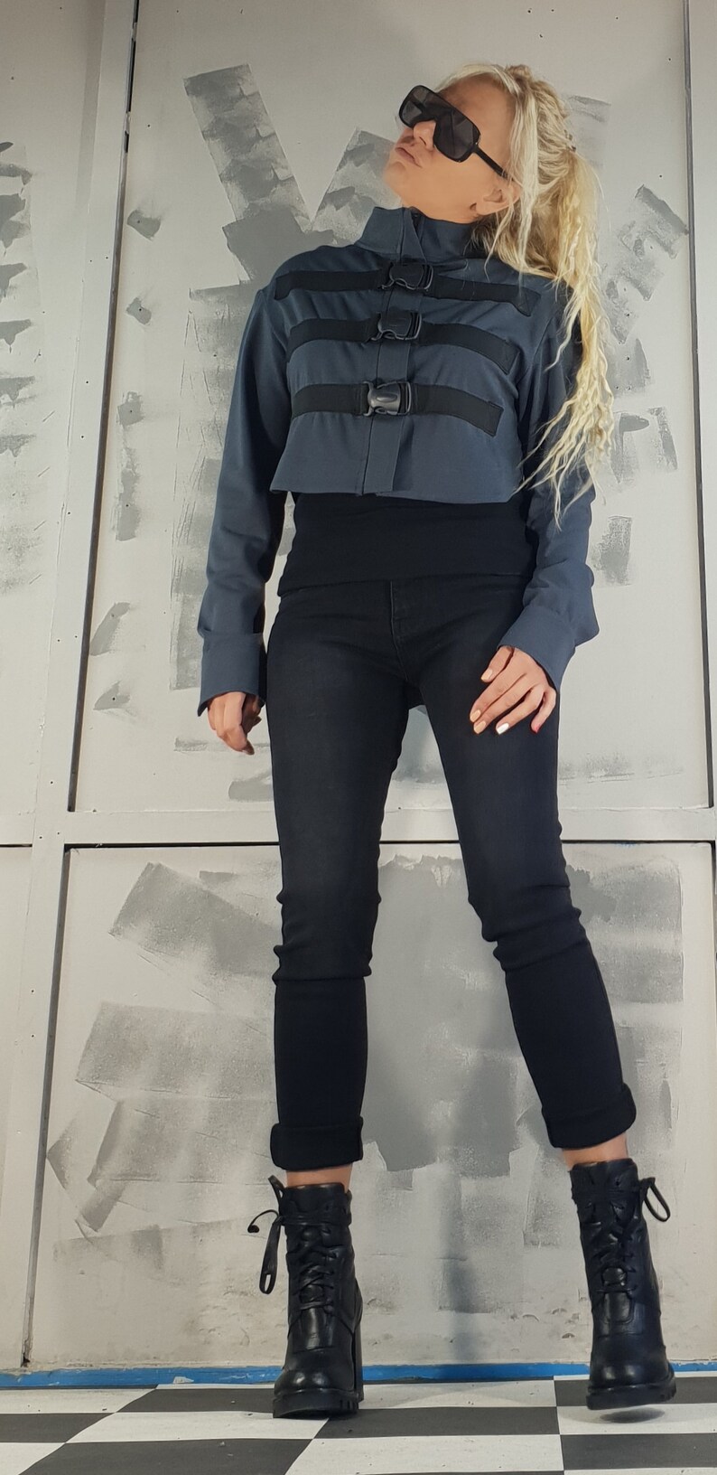 May include: A woman wearing a blue cropped jacket with black straps and buckles, black jeans, and black lace-up boots. She is wearing sunglasses and has blonde hair.