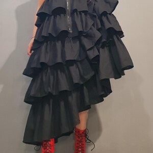 May include: A black sleeveless dress with multiple layers of ruffles. The dress is asymmetrical, with one side longer than the other. The model is wearing red lace-up boots.