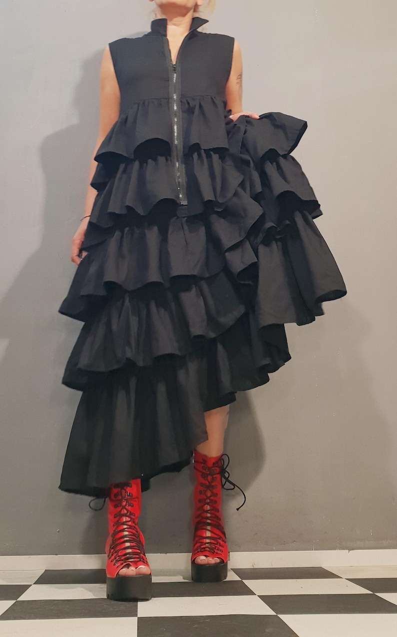 May include: A black sleeveless dress with multiple layers of ruffles. The dress is asymmetrical, with one side longer than the other. The model is wearing red lace-up boots.