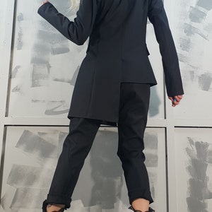 May include: A black blazer with a long, asymmetrical hemline, paired with black pants and black boots. The person is standing in front of a wall with gray paint.