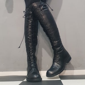 May include: Black leather knee-high boots with lace-up detailing. The boots have a flat sole and a sleek, streamlined design. The laces extend up the front of the boots, adding a touch of style. The boots are shown against a gray and black checkered floor.
