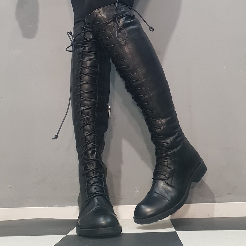 May include: Black leather knee-high boots with lace-up detailing. The boots have a flat sole and a sleek, streamlined design. The laces extend up the front of the boots, adding a touch of style. The boots are shown against a gray and black checkered floor.