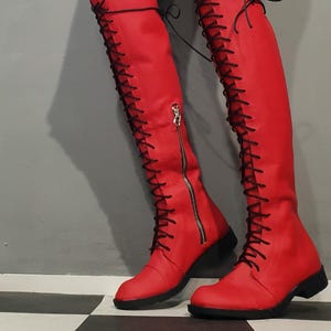 May include: Red knee-high boots with black laces and a side zipper. The boots have a low heel and a rounded toe. The laces run up the front of the boots. The boots are made of a smooth material.