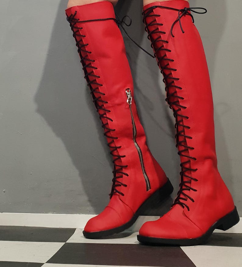 May include: Red knee-high boots with black laces and a side zipper. The boots have a low heel and a rounded toe. The laces run up the front of the boots. The boots are made of a smooth material.