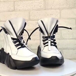 May include: A pair of white high-top boots with black accents. The boots feature a chunky black sole, a zipper down the front, and black laces. The boots are displayed on a white surface.