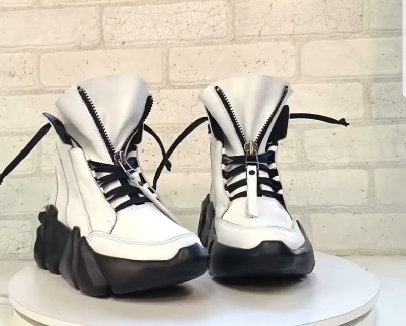 May include: A pair of white high-top boots with black accents. The boots feature a chunky black sole, a zipper down the front, and black laces. The boots are displayed on a white surface.